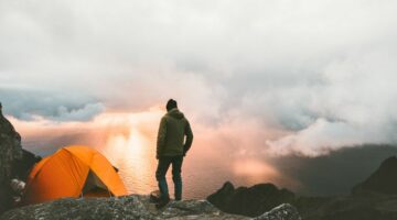 Mann steht neben Zelt auf dem Berg und schaut Sonnenuntergang an
