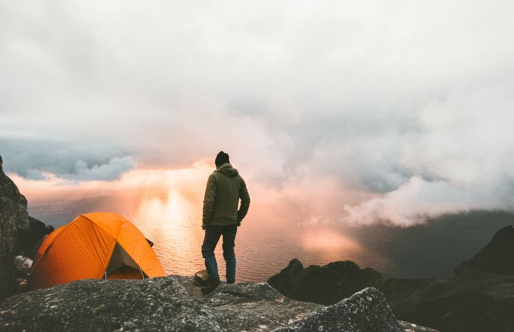 Mann steht neben Zelt auf dem Berg und schaut Sonnenuntergang an
