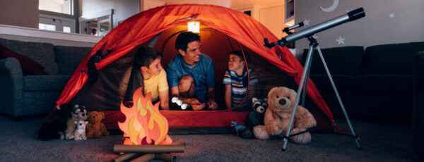 A dad with his two sons camp inside their home due to the coronavirus restrictions and quarantine. They have pitched a tent along with stuffed animal friends and have a fake campfire next to a telescope. They are making the best of their situation and long to return to the outdoors.